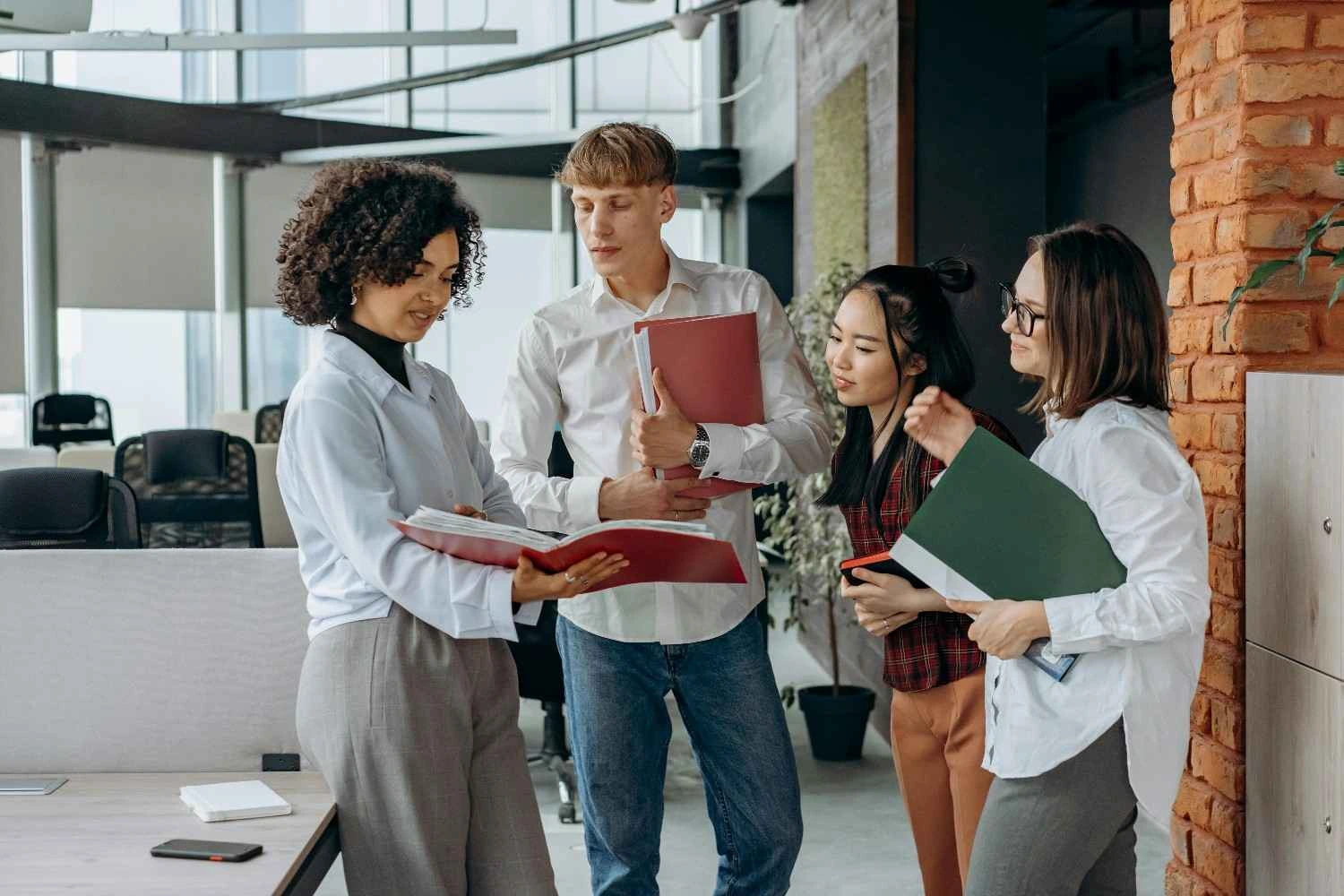 Empat rekan kerja dalam magang yang berkeinginan menjadi karyawan tetap dari berbagai latar belakang sedang berdiskusi di kantor modern yang bergaya industrial. Seorang wanita berambut ikal memegang map merah terbuka, sementara rekan-rekannya mendengarkan dan memperhatikan dokumen tersebut di area kerja terbuka dengan dinding bata ekspos.