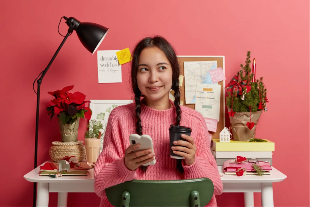 Seorang remote worker memegang smartphone dan kopi sambil bekerja di meja kerja yang rapi, menggambarkan pentingnya membangun personal branding sebagai remote worker melalui kehadiran profesional di dunia digital.