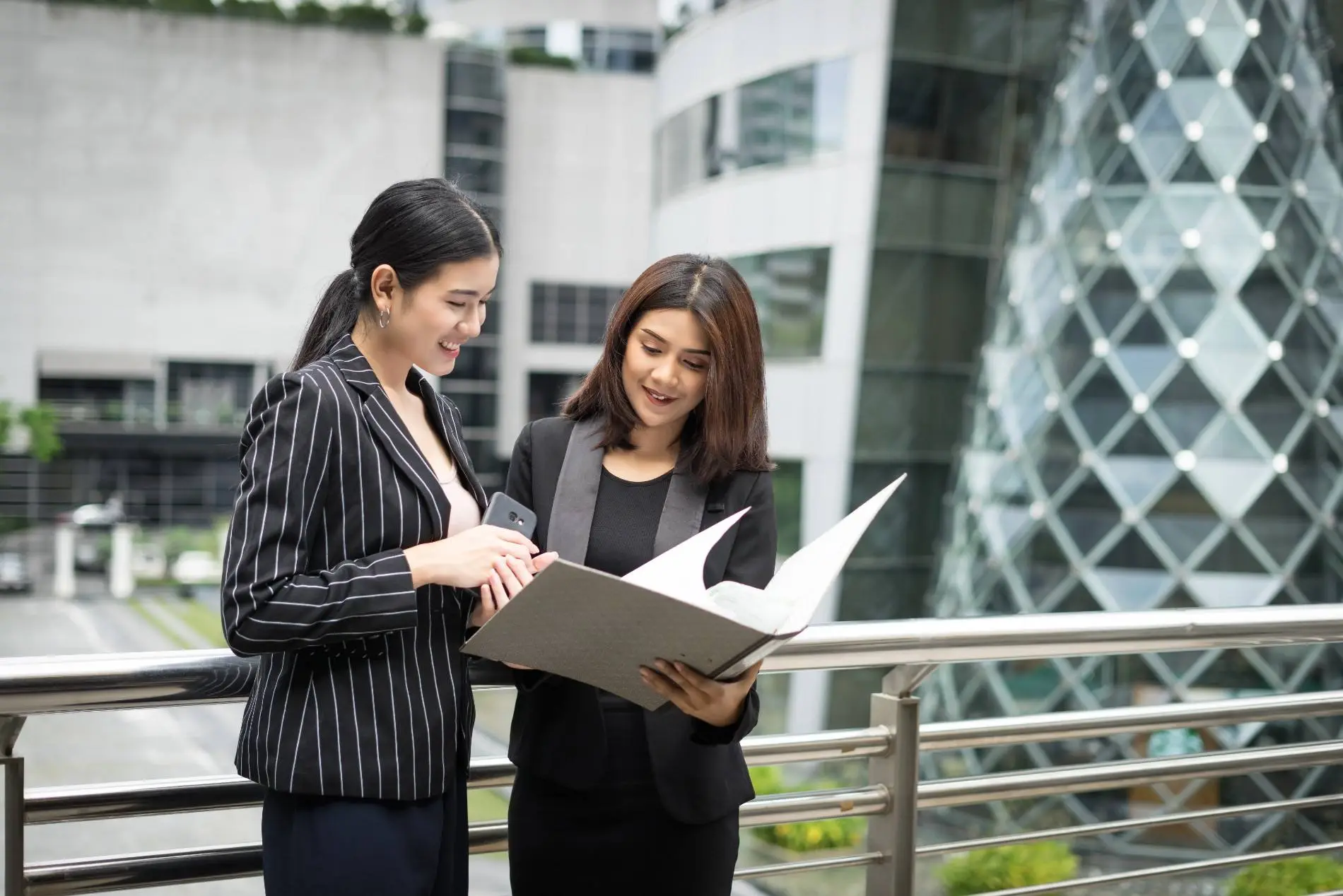 Dua profesional sedang berdiskusi sambil meninjau dokumen di depan gedung perkantoran, menggambarkan persiapan dan strategi sebagai bagian dari cara lolos seleksi di perusahaan asing